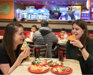 Two women sit at a table, each has a plate of pizza slices and a plate of salad. They are smiling as they each eat their own slice of pizza.
