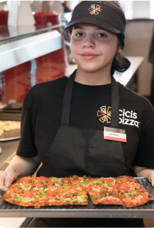 An employee stands next to a Cicis buffet line, holding a freshly baked supreme pizza. She wears an apron, black tee shirt, with the Cicis pizza spark logo and text reading 'Cicis Pizza', and a black visor with the Cicis logo.
