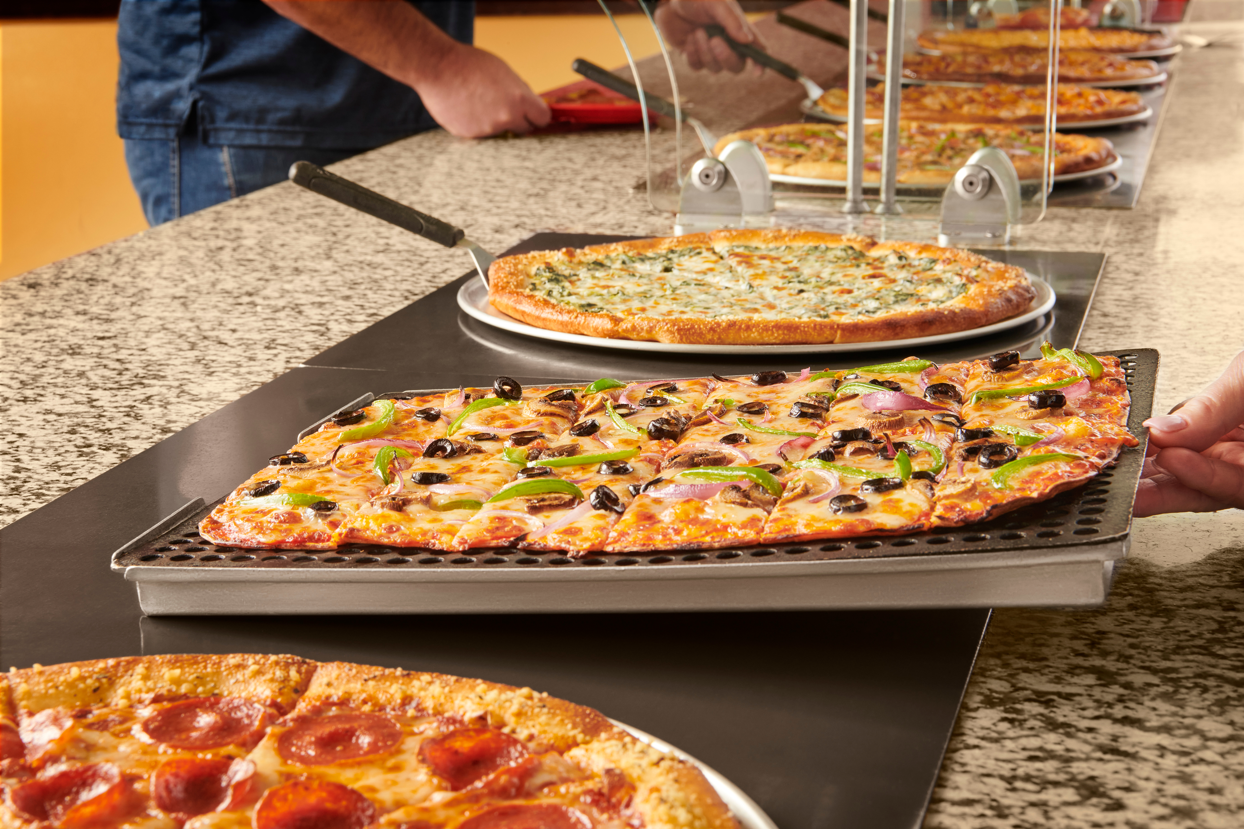 View down a full Cicis pizza buffet line. A customer is lifting a pizza slice to add to their plate.