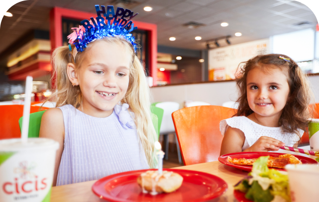 Two young girls sit next to each other at a table in a Cicis Pizza Buffet. One girl is wearing a crown that says "Happy Birthday" and has a Cicis cinnamon roll topped with lit birthday candle in front of her.