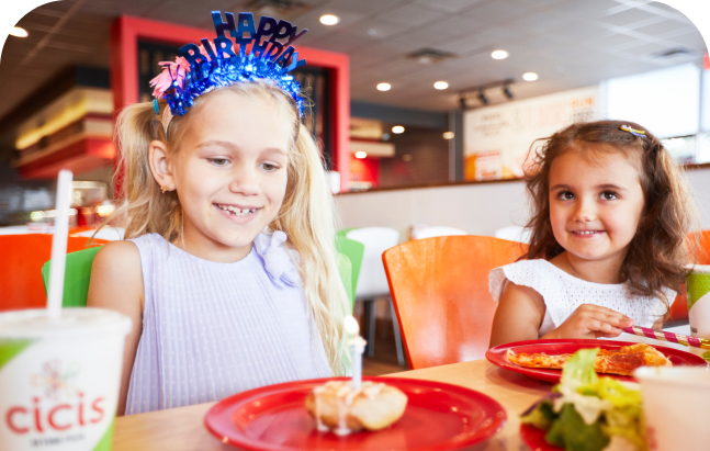 Two young girls sit next to each other at a table in a Cicis Pizza Buffet. One girl is wearing a crown that says "Happy Birthday" and has a Cicis cinnamon roll topped with lit birthday candle in front of her.