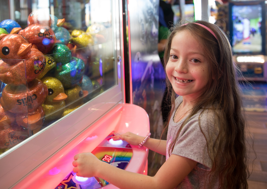 A young girl smiles as she plays an arcade game to win a prize. The arcade machine is filled with colorful rubber ducks with "Cicis" written on each one.